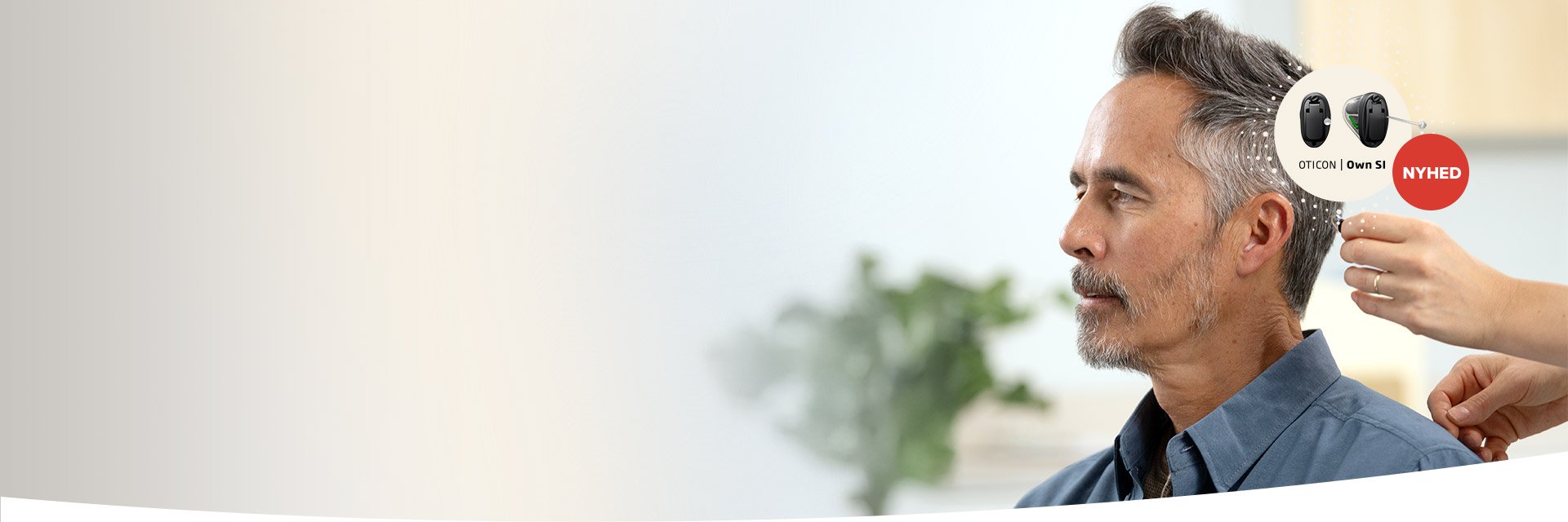 En mand iført blå skjorte får tilpasset et høreapparat af en audiolog. Tekst: ’OTICON | Own SI’ og ’NYHED’. Baggrunden viser en indendørs plante og hvide vægge.