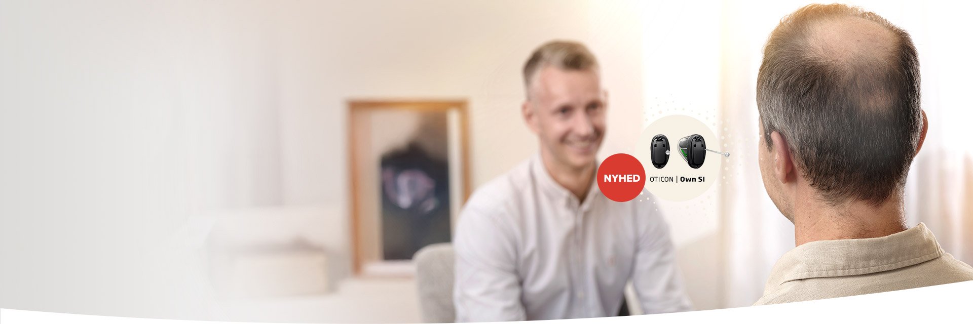 En mand med rygvendt perspektiv sidder overfor en audiolog i en høreklinik. På billedet vises høreapparater fra Oticon med teksten ’NYHED OTICON | Own S1’.