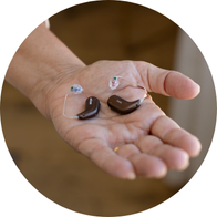 En hånd holder to diskrete høreapparater med transparent ledning og ørepropper. Baggrunden er sløret, sandsynligvis i en høreklinik som Audika, der tilbyder hørepleje og høretests.