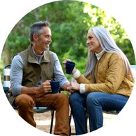 To ældre personer sidder udendørs på stole og drikker kaffe fra blå krus, mens de holder hinandens hænder. Baggrunden viser grøn natur med træer og lysindfald. Ingen tekst identificeret.