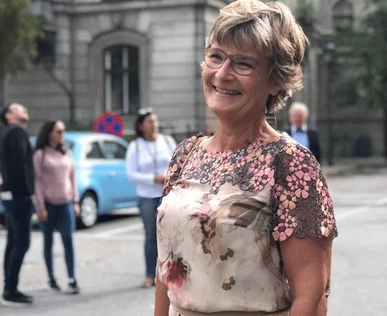 En ældre kvinde iført blomstret bluse står udendørs i en bymæssig gade med mennesker og biler i baggrunden ved en historisk bygning med buede vinduer.