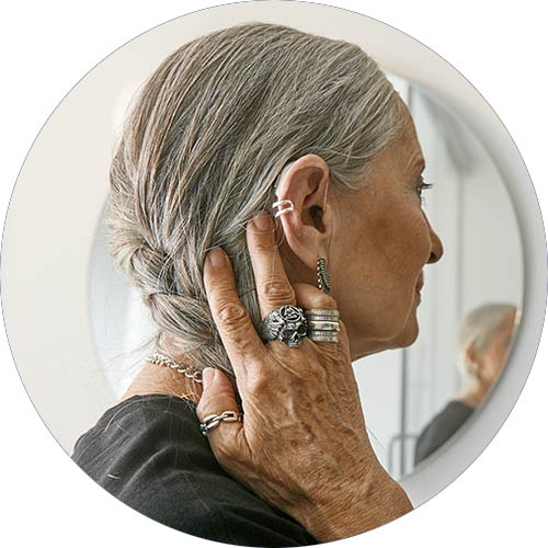 En ældre kvinde med gråt hår justerer en diskret høreapparat bag øret med hånden. I baggrunden ses et spejl, der reflekterer dele af en klinikindretning.