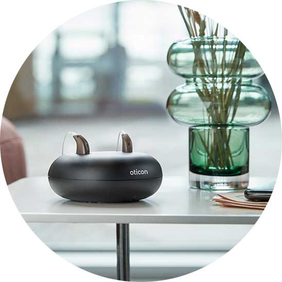 Trådløs oplader til Oticon høreapparater placeret på et bord ved siden af en moderne glasvase og en smartphone
