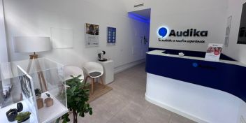 Recepción de una clínica auditiva Audika; mostrador azul con logo ’Audika: Tu audición al cuidado de especialistas’, audífonos exhibidos en vitrina, folletos informativos y asientos en ambiente moderno y luminoso.