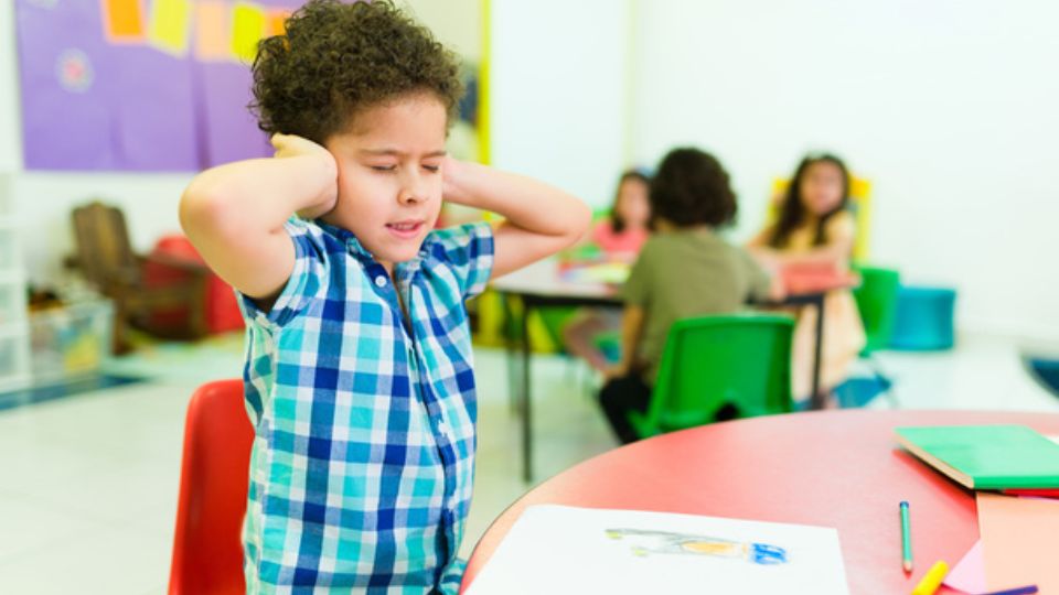niño en el aula cubriéndose los oídos mientras otros niños realizan actividades.