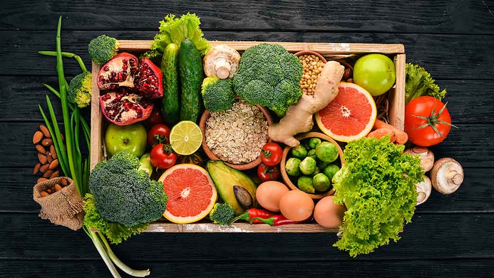 Una caja de madera contiene alimentos saludables como brócoli, granada, huevos, pomelos, aguacates, avena, lechuga y tomate. Todo está dispuesto sobre una mesa negra.