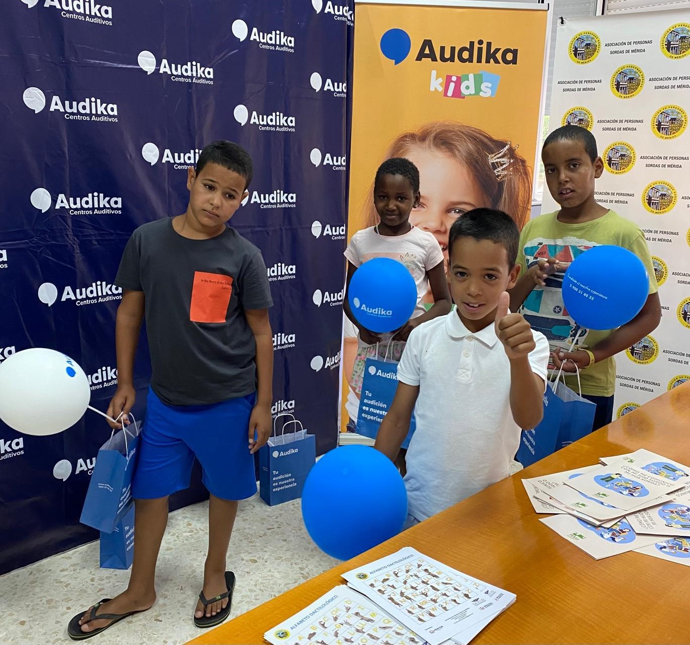 Niños participando en una actividad solidaria organizada por Audika, con globos y bolsas promocionales en un entorno decorado con pancartas de la marca.