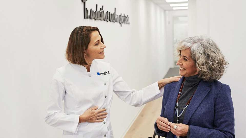 Una audioprotesista con bata blanca de Audika atiende amablemente a una mujer mayor de cabello canoso en un pasillo iluminado de una clínica auditiva. En la pared se lee: ’Tu audición es nuestra prioridad’.
