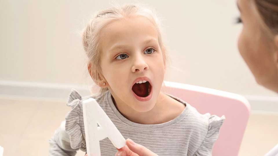 Una niña sostiene una letra ’A’ blanca mientras está sentada en una silla rosa en un entorno interior iluminado, con una pared beige y suelo claro en el fondo.