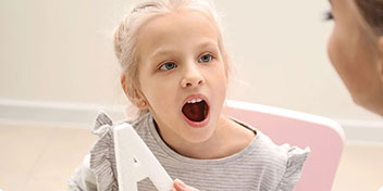 Una niña sujetando una letra ’A’ blanca de madera, sentada en una silla rosada. El fondo es claro y minimalista, con paredes beige y un piso brillante.