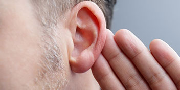 Un hombre coloca su mano detrás de su oreja derecha en un gesto de ’escuchar mejor,’ con fondo gris neutro, evocando temas de audición y cuidado auditivo especializado.
