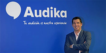 Un hombre de brazos cruzados, con rostro difuminado, está de pie frente a una pared azul con el logo de Audika y el texto: ’Tu audición es nuestra experiencia’.