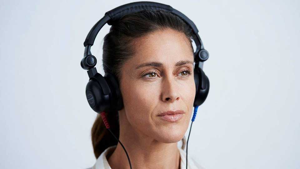 Auriculares colocados sobre la cabeza de una persona con rostro difuminado, utilizados posiblemente para una prueba de audición. Fondo blanco minimalista típico de una clínica auditiva.