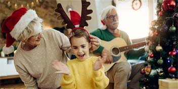 niña con diadema de reno bailando junto a su abuela con gorro de Navidad mientras un hombre toca la guitarra cerca del árbol de Navidad
