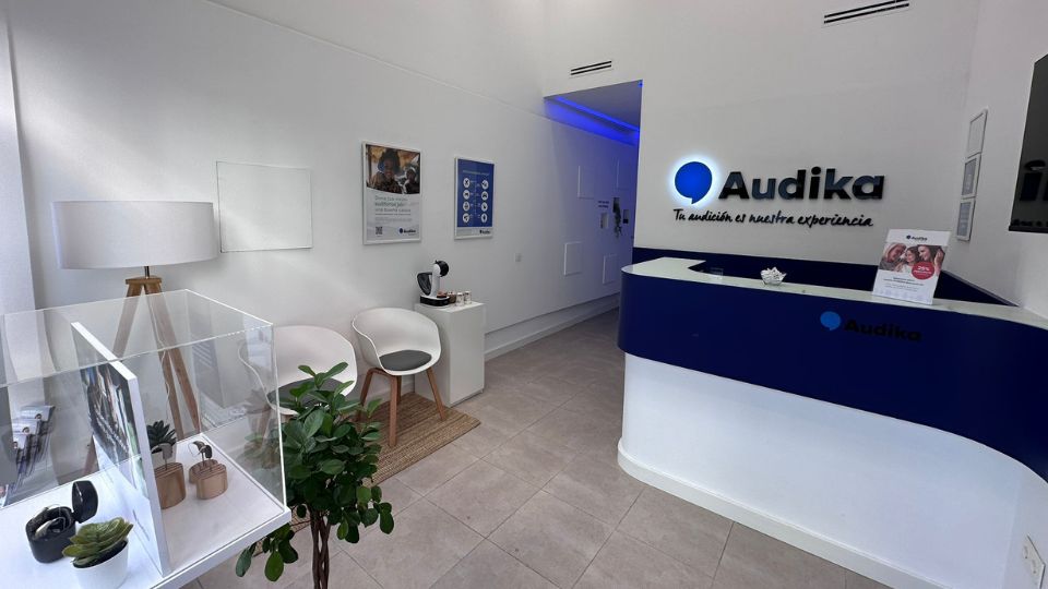 Mostrador azul de Audika con el lema ’Tu audición es nuestra experiencia’ en una clínica moderna. A la izquierda, hay sillas blancas, plantas decorativas y una vitrina con audífonos.
