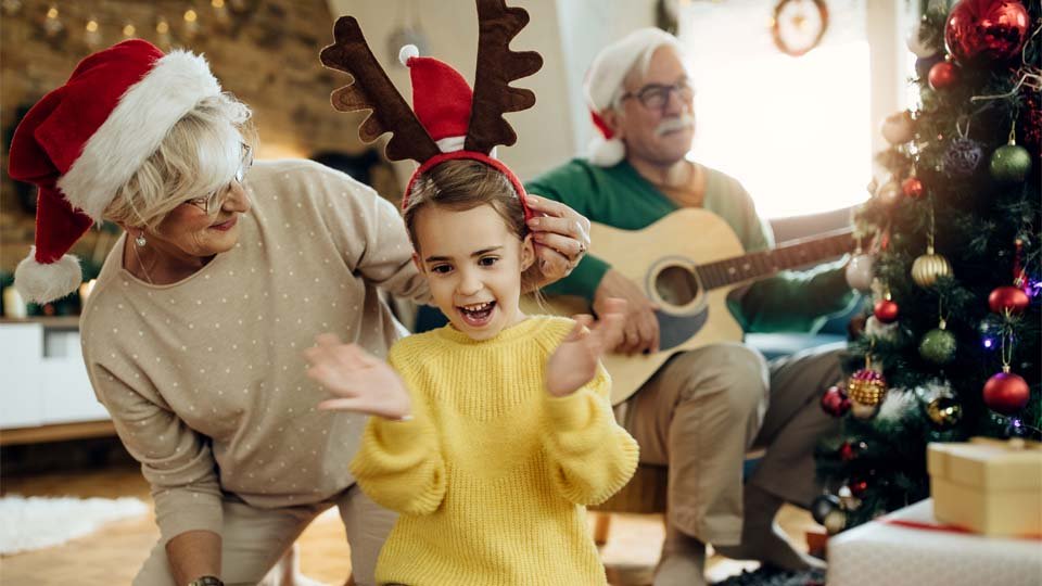 Niña con diadema de reno y abuela con gorro de Navidad disfrutando juntas mientras un hombre toca la guitarra cerca del árbol decorado.