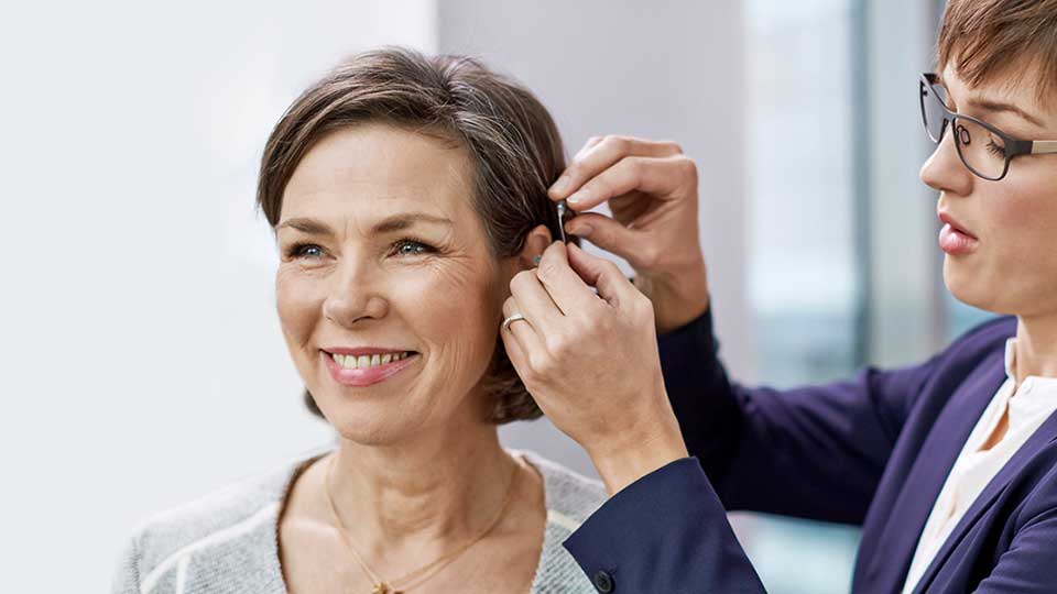 Una audioprotesista ajusta un audífono detrás de la oreja de una mujer en una consulta en una clínica auditiva, con fondo iluminado por ventanas grandes.