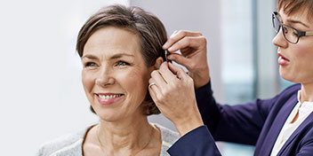 Un audiólogo ajusta un audífono detrás de la oreja de una mujer en un entorno clínico bien iluminado. Podría tratarse de una consulta en una clínica auditiva especializada como Audika.