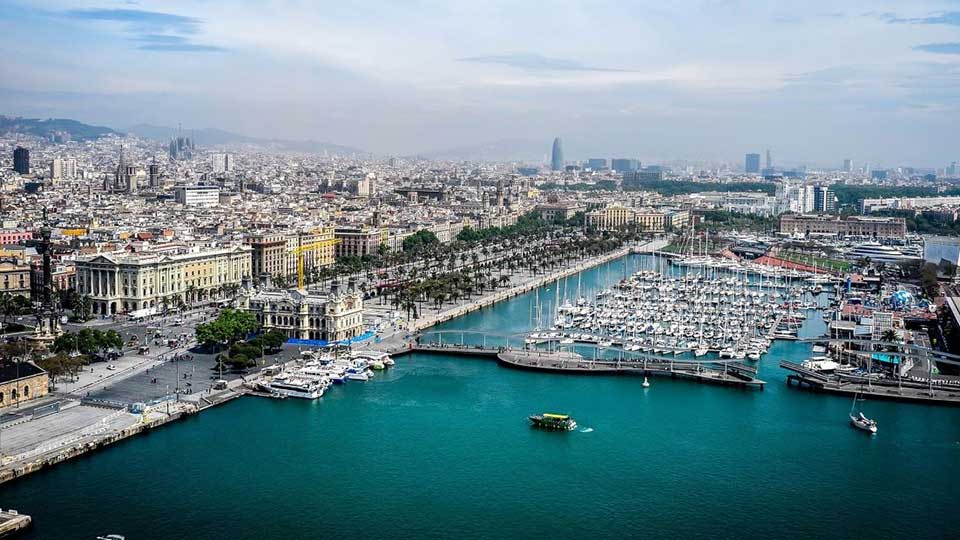Un puerto marítimo lleno de yates está rodeado por edificios históricos y palmeras en Barcelona; al fondo se destacan la Sagrada Familia y la Torre Glòries bajo un cielo despejado.
