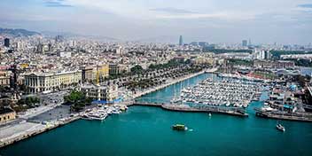 Vista panorámica del puerto de Barcelona, donde yates y barcos están atracados en el muelle; la ciudad se extiende al fondo con edificios emblemáticos y cielo despejado.