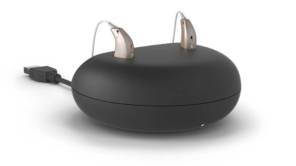 Dos audífonos beige tipo receptor en canal están sobre un cargador negro con diseño ovalado, conectado por un cable USB; pequeñas luces verdes indican actividad. Contexto: atención auditiva.