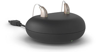 Dos audífonos recargables descansan en su base de carga negra conectada por un cable USB. Luces verdes indican carga activa en un entorno minimalista y limpio.