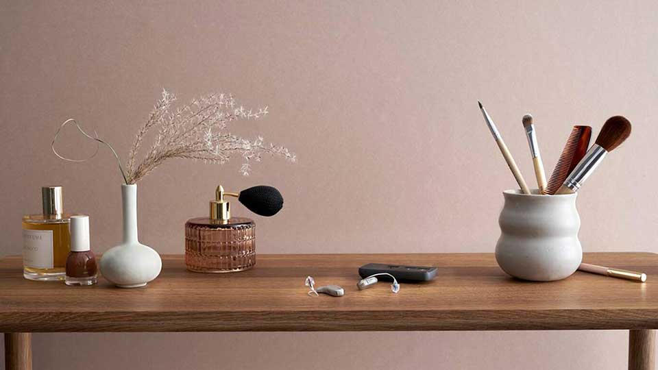Audífonos discretos descansan sobre una mesa de madera junto a perfumes, pinceles y accesorios de maquillaje en un ambiente minimalista con fondo beige, representando cuidado y bienestar personal.