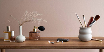 Dos audífonos discretos están colocados sobre una elegante mesa de madera junto a objetos decorativos y de belleza como perfumes, pinceles y un jarrón, en un ambiente minimalista.