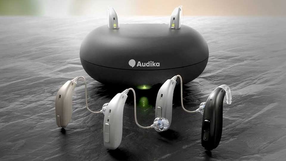 Un cargador negro de Audika contiene cuatro audífonos de distintos colores (beige, blanco, plateado y negro) colocados alrededor, sobre una superficie gris, en un ambiente iluminado.