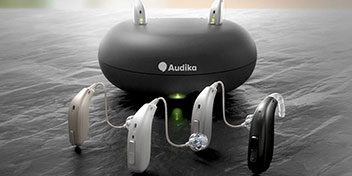 Cuatro audífonos modernos en diferentes colores (beige, blanco, plateado y negro) están alineados frente a un cargador negro de Audika, sobre una superficie oscura en un entorno iluminado.