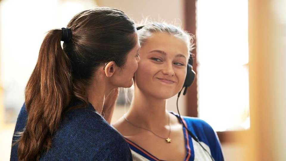 Una mujer con auriculares realiza una prueba de audición en un entorno iluminado con luz natural, mientras otra persona cercana parece asistirla o hablarle suavemente. No hay texto visible.