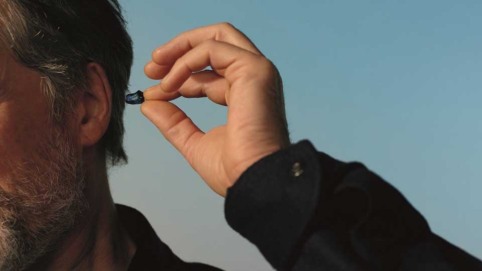 Un hombre sostiene y coloca un audífono intracanicular azul en su oído. Fondo azul claro indica un ambiente al aire libre. Ideal para servicios de cuidado auditivo en Audika.