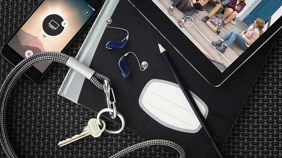 Dos audífonos azules de Audika reposan sobre un cuaderno junto a un lápiz, una tableta mostrando un video, un móvil conectado a TV y llaves sobre superficie negra tejida.