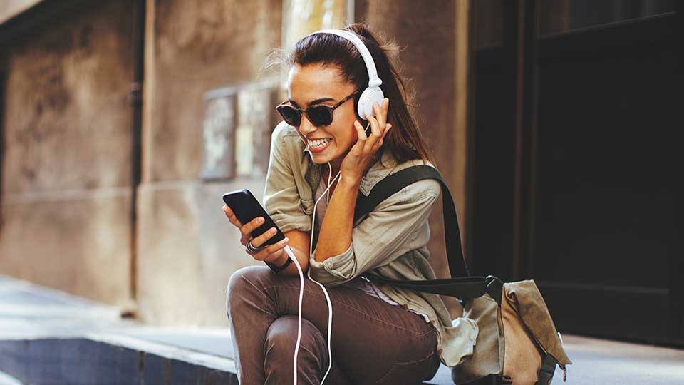 Una mujer con auriculares blancos escucha música desde su teléfono móvil mientras está sentada en la acera, rodeada de un entorno urbano con edificios de fondo.