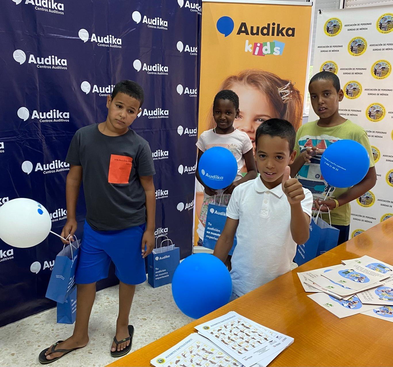 Niños participando en una actividad solidaria organizada por Audika, con globos y bolsas promocionales en un entorno decorado con pancartas de la marca.