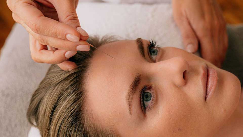 Manos ajustan cuidadosa y delicadamente un dispositivo cerca del oído de una persona con cabello canoso sobre una superficie acolchada blanca, posiblemente en un entorno de clínica auditiva. No hay texto visible.