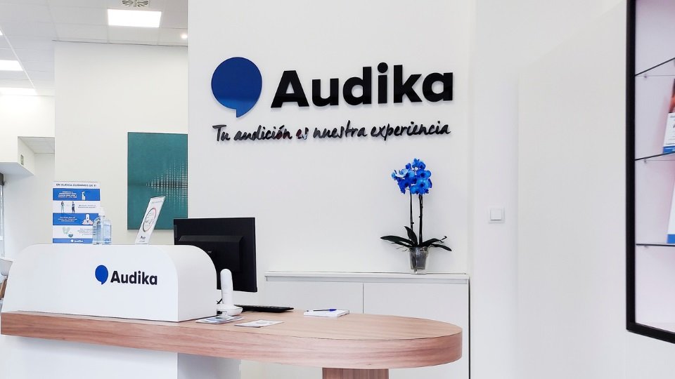 Recepción del centro auditivo Audika en Menorca, con mostrador blanco, detalles minimalistas, logotipo y eslogan en la pared, y decoración con orquídea azul.