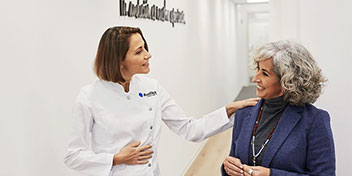 Una audioprotesista con uniforme blanco de Audika interactúa amablemente con una mujer mayor de cabello canoso en un pasillo blanco de una clínica auditiva. Texto visible: ’We believe sound connects us.’