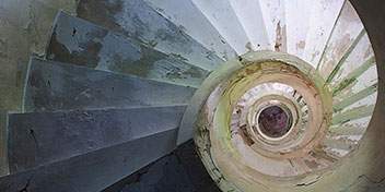 Una escalera de caracol desgastada gira hacia abajo, con peldaños de concreto dañados y paredes marcadas por humedad y moho, creando un ambiente envejecido y abandonado.