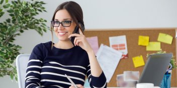 mujer joven hablando por teléfono mientras sostiene un lápiz en una oficina con un tablero de corcho y un portátil sobre el escritorio