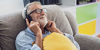 Un hombre mayor usa auriculares sobre la cabeza mientras se reclina en un sofá gris, sosteniendo un cojín amarillo. Al fondo, hay muebles coloridos en un entorno doméstico iluminado.