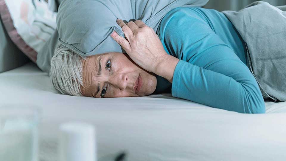 Una persona mayor con cabello canoso, acostada en una cama cubierta con sábanas grises, se cubre los oídos con una almohada en una habitación iluminada. No hay texto visible.