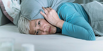 Una persona mayor recostada en una cama sostiene una almohada contra su cabeza. Lleva ropa azul y está en un entorno doméstico con frascos desenfocados en primer plano.