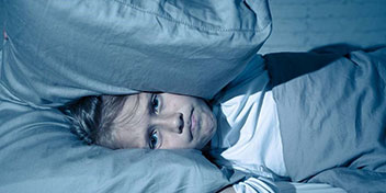 Un niño aparece acostado en una cama con expresión preocupada, rodeado de almohadas grises en un ambiente nocturno con luz tenue, transmitiendo incomodidad o insomnio. No hay texto visible.
