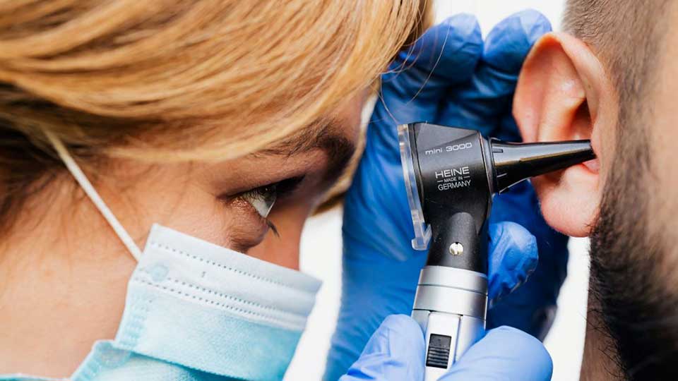 Un otoscopio Heine mini 3000 está siendo utilizado por un audioprotesista con guantes azules para examinar el oído de un paciente masculino en un entorno clínico especializado en salud auditiva.