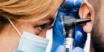 Un audiólogo con guantes azules examina el oído de un hombre usando un otoscopio en una clínica auditiva. El entorno sugiere un procedimiento de revisión para un test de audición.