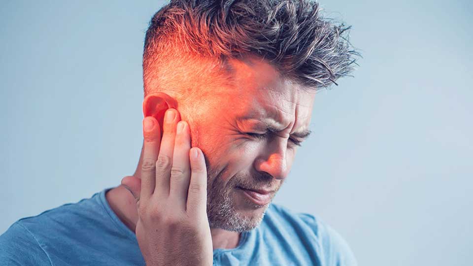 Un hombre con cabello corto toca su oreja izquierda con expresión de molestia, destacando un área iluminada en rojo para simbolizar dolor o problema auditivo; fondo azul claro neutro.