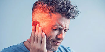 Un hombre toca su oído izquierdo con expresión de incomodidad (rostro difuminado); fondo azul claro. Podría sugerir preocupación por pérdida auditiva o necesidad de consulta en una clínica auditiva.