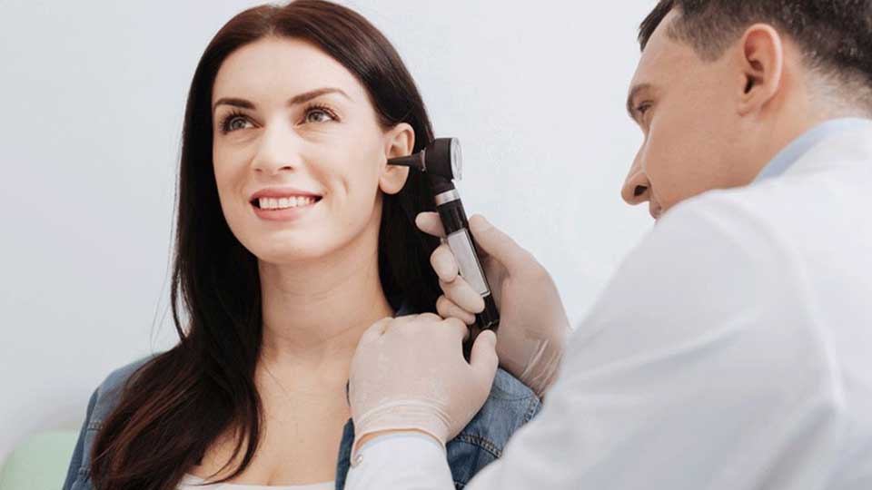 Un audiólogo utiliza un otoscopio para examinar el oído de una mujer en una clínica auditiva. La paciente lleva el cabello suelto, y el ambiente es profesional y luminoso.