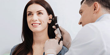 Un audiólogo, usando guantes, realiza un examen auditivo con un otoscopio en el oído de una mujer en una clínica especializada en cuidado auditivo. Fondo blanco, ambiente profesional.